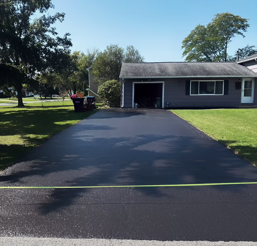 Freshly sealcoated driveway leading to a gray ranch-style home with green lawn and mature trees on a sunny day in Rochester NY