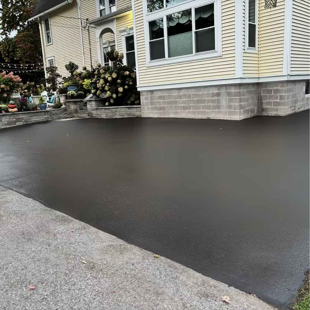 Freshly sealcoated residential driveway in front of a yellow sided home with stone retaining walls and landscaping in Rochester NY